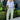 Woman wearing a light blue top and green patterned pants standing outdoors.