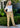 Woman wearing a white top and colorful pants standing outdoors with greenery around.