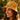 A woman wearing a yellow patterned bucket hat with red and green floral motifs, photographed outdoors against a warm, sunlit green background.