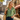 A woman walking on a cobblestone street wearing a green patterned blouse and dark blue jeans, carrying a woven straw tote bag. In the background are classic European buildings and a cafe with a red awning.